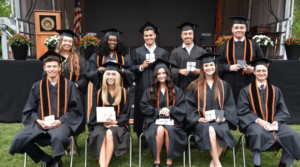 Studenti e studentesse diplomati della The Linsly School in abito accademico davanti a un edificio con la bandiera americana.