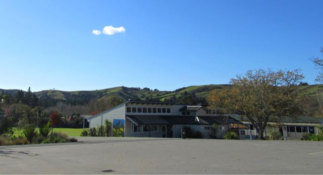 Das weiße Gebäude von Kaipara College steht eingebettet zwischen sanften Hügeln und grüner Landschaft unter blauem Himmel.