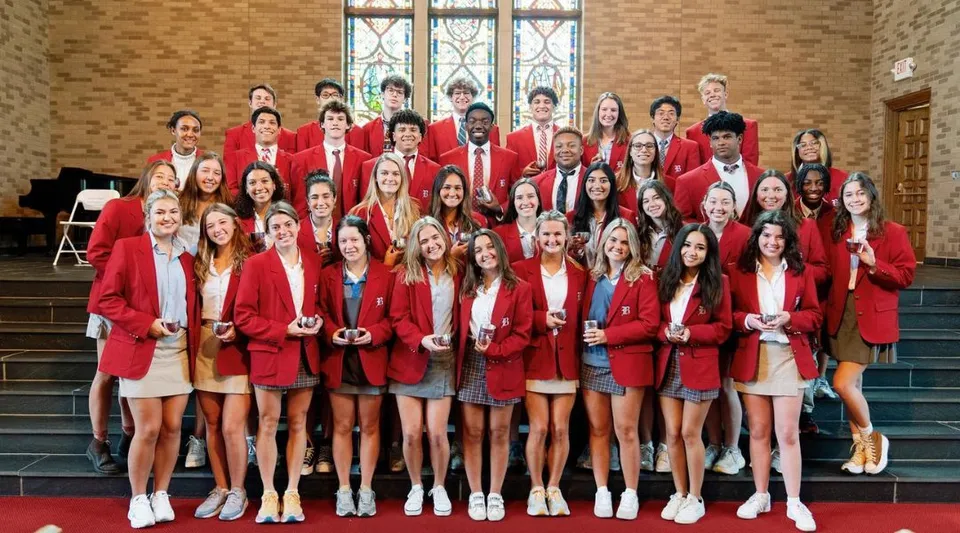 Un gruppo di studenti e studentesse della Baylor School in toga rossa sta sulle scale davanti a un edificio con vetrate colorate.