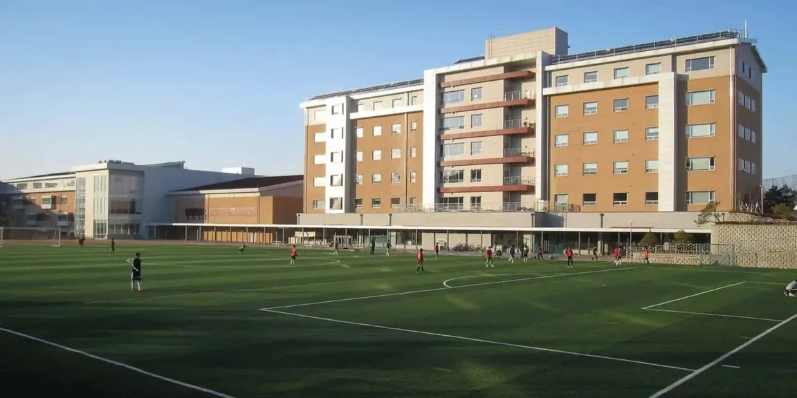 Schulgebäude mit mehrstöckigem Wohnheim hinter einem Sportplatz, auf dem Schülerinnen und Schüler Fußball spielen