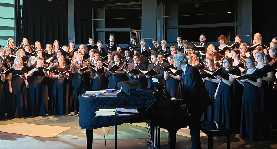 Il coro della Holy Family Catholic High School si trova sul palco con tuniche nere e blu davanti a un pianoforte a coda.