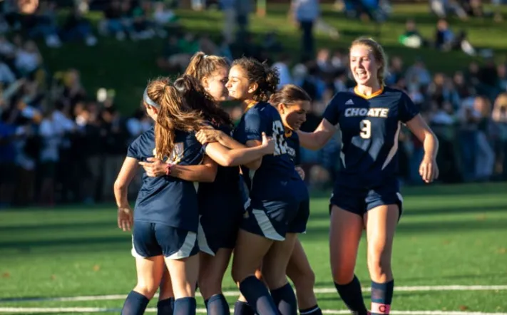 Un grupo de jóvenes futbolistas de Choate Rosemary Hall celebra juntas en un campo de césped ante espectadores.