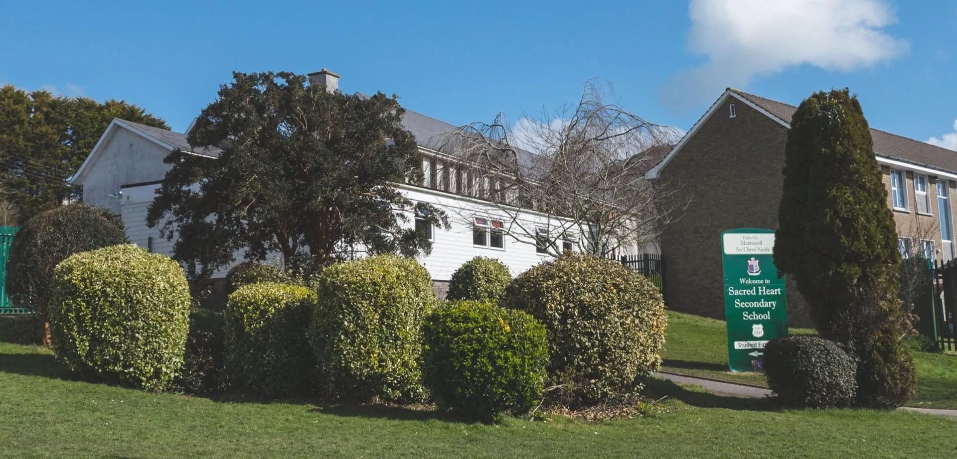 L'edificio storico della Sacred Heart Secondary School Clonakilty circondato da siepi ben curate e alberi sotto un cielo azzurro limpido.