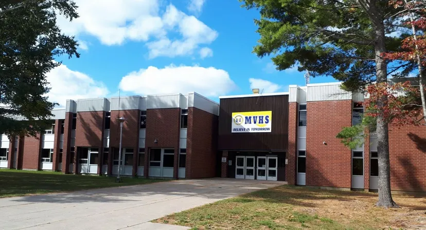 L'edificio in mattoni della Miramichi Valley High School è circondato da alberi e un prato sotto un cielo azzurro.