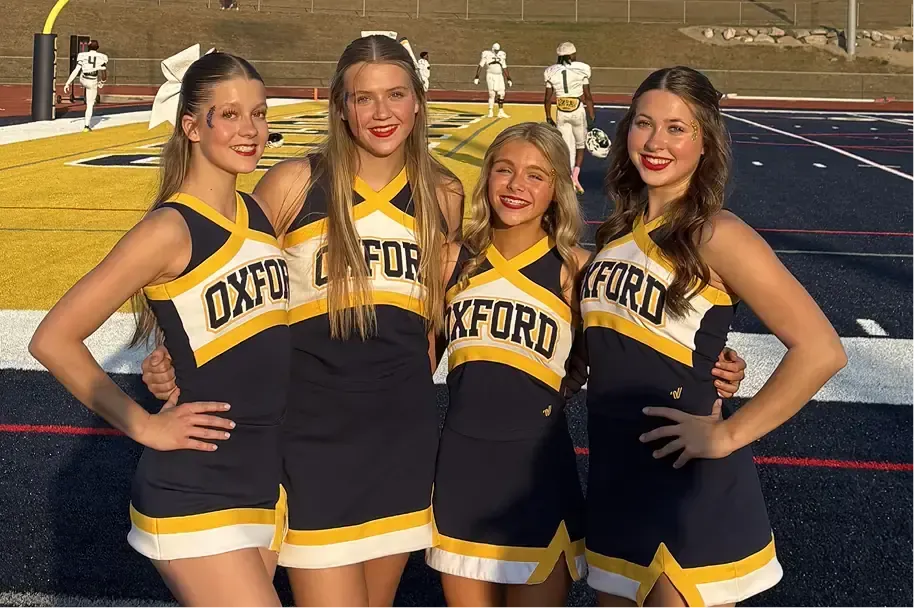 Cuatro animadoras con uniforme de Oxford sonríen junto al campo de fútbol americano