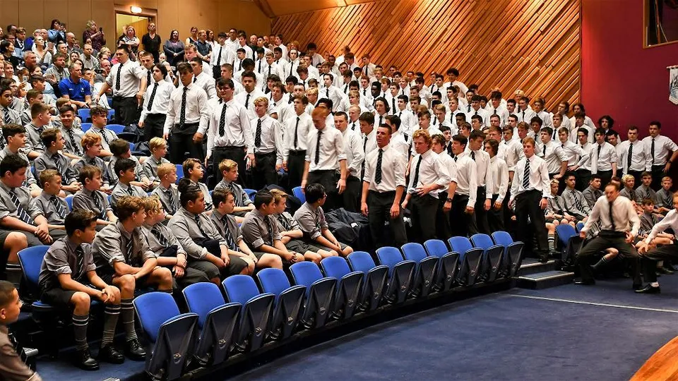 Un gruppo di studenti è seduto su sedie blu in una stanza con pareti in legno nel campus della Otago Boys' High School.