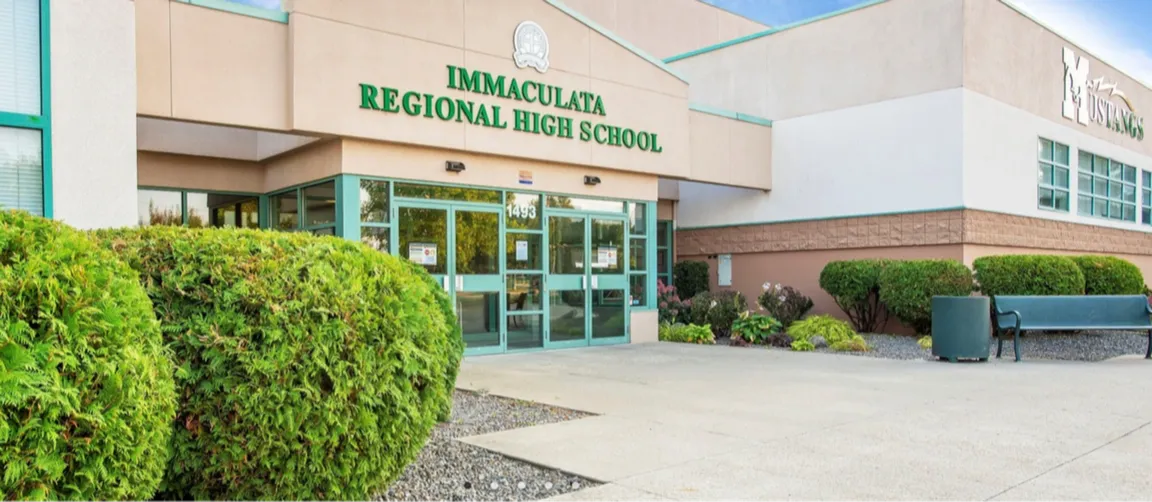 Eingangsbereich eines Schulgebäudes mit Schriftzug Immaculata Regional High School, Sträuchern und Sitzbank vor dem Eingang