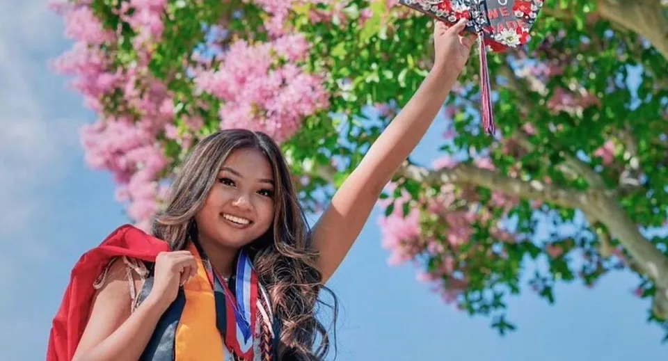 Una giovane donna con lunghi capelli scuri alza le braccia sorridendo, circondata da fiori rosa nel campus dell'Anaheim Union High School District.