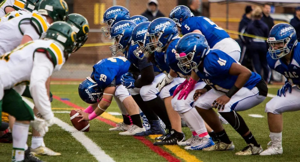 Studenti del John Abbott College in maglia blu e verde sul campo di gioco si preparano a un'azione di football.