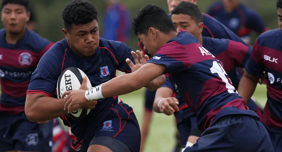 Due giocatori si contendono la palla durante una partita di rugby sul campo del Macleans College, mentre altri studenti si trovano sullo sfondo.
