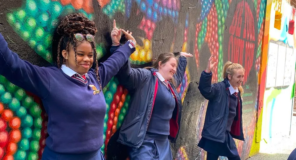Un gruppo di studenti e studentesse della Sacred Heart Secondary School in divisa scolastica si trova davanti a un murale colorato e alza le braccia.