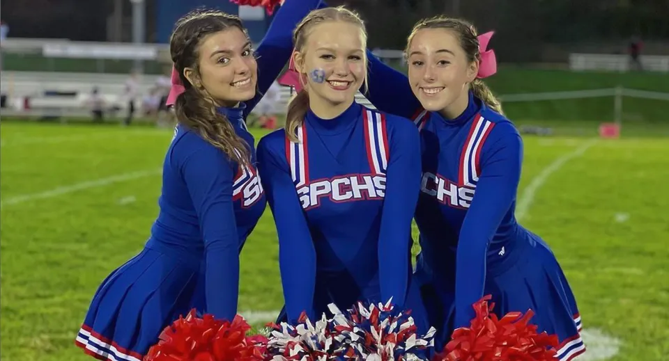 Tre studentesse della St. Paul Catholic High School posano in divisa da cheerleader con i pompon sul campo sportivo di notte.