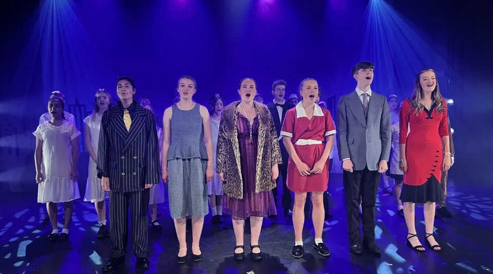 Un gruppo di studenti e studentesse della St. Dominic's Grammar School presenta una sfilata di moda su un palco con illuminazione blu e viola.