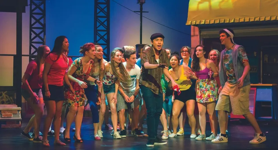 Un gruppo di studenti in costumi colorati si trova sul palco della Brentwood College School davanti a luci e attrezzature di scena.