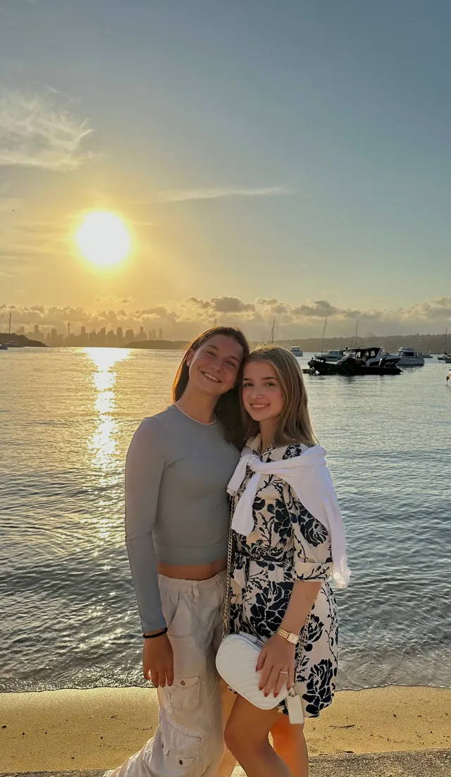 Dos chicas posando en la orilla al atardecer con veleros fondeados en la bahía