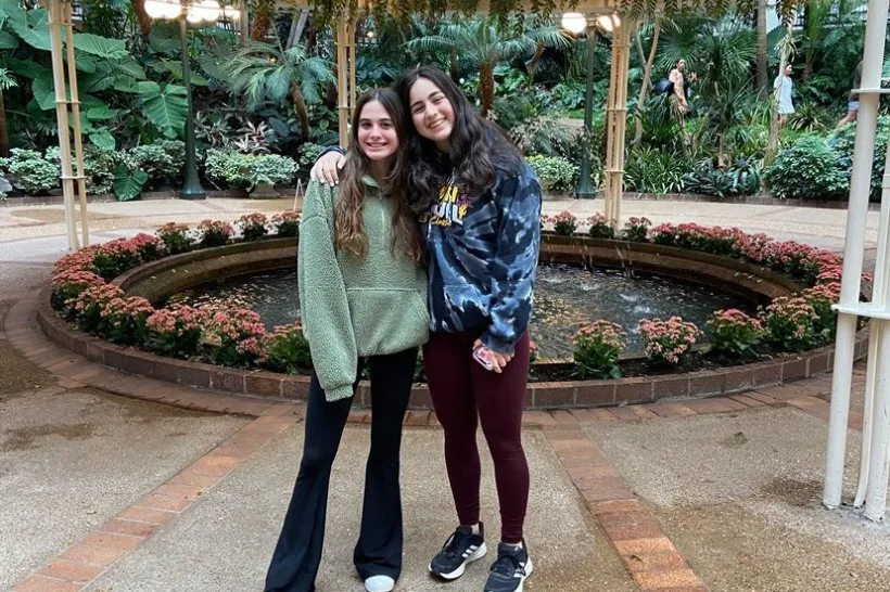 Dos chicas jóvenes sonrientes posan abrazadas junto a una fuente circular en un jardín interior