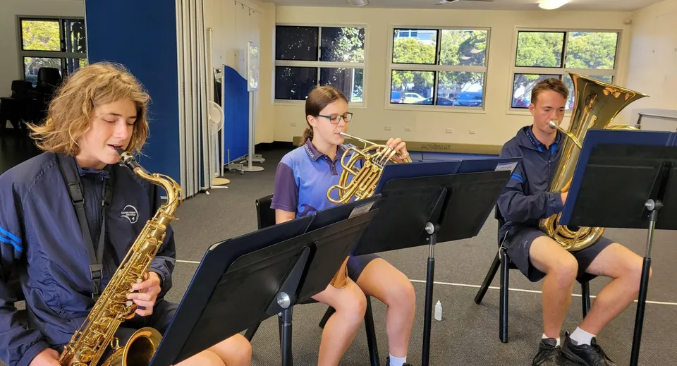 Studenti del Kawana Waters State College suonano strumenti come sassofono, trombone e tuba in un'aula di musica.