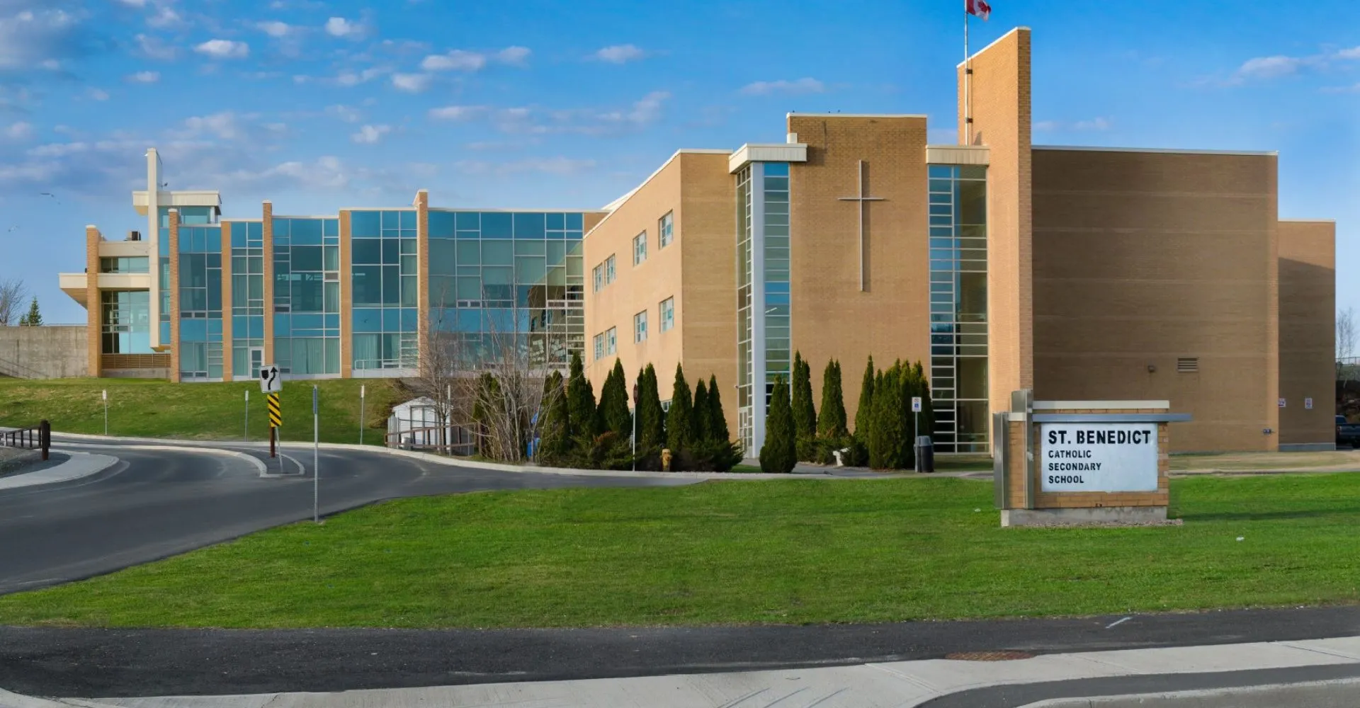 L'edificio scolastico moderno a più piani della St. Benedict Catholic Secondary School con facciata in vetro e giardino curato sotto un cielo blu.