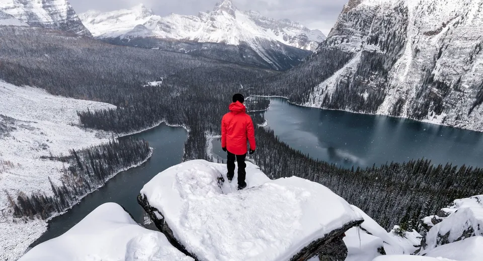 Una persona con una giacca rossa si trova su una scogliera innevata con vista su un lago ghiacciato e montagne coperte di neve presso la St. Pius X High School.