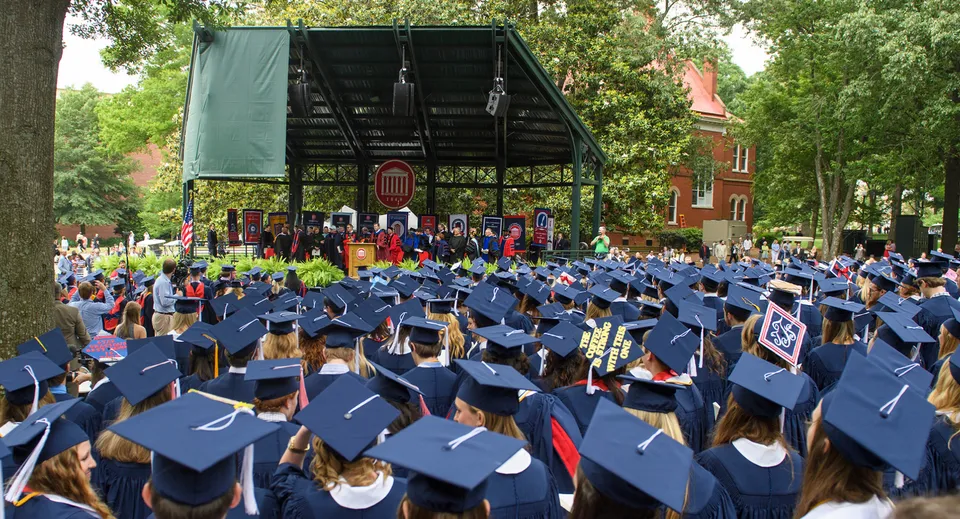 I diplomati della Sidcot School si radunano all'aperto in toghe e tocchi blu durante la cerimonia di diploma davanti a un palco.