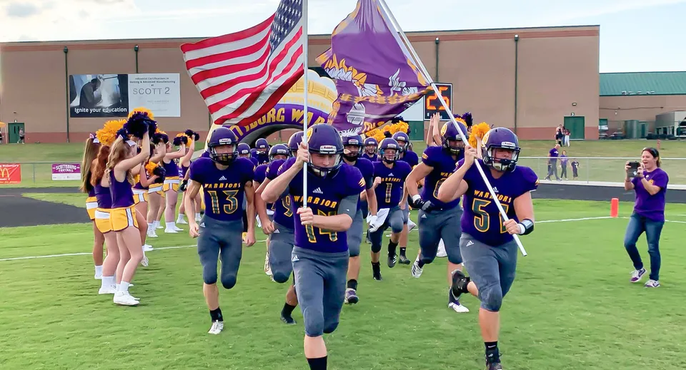 Un gruppo di giocatori di football in uniformi viola porta bandiere ed entra in campo nel campus della Scottsburg High School.
