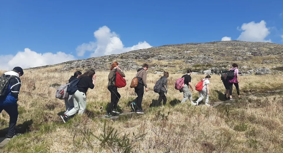 Un gruppo di studenti del Coláiste Muire Crosshaven fa un'escursione su un sentiero erboso in un paesaggio montuoso sotto un cielo blu con nuvole.