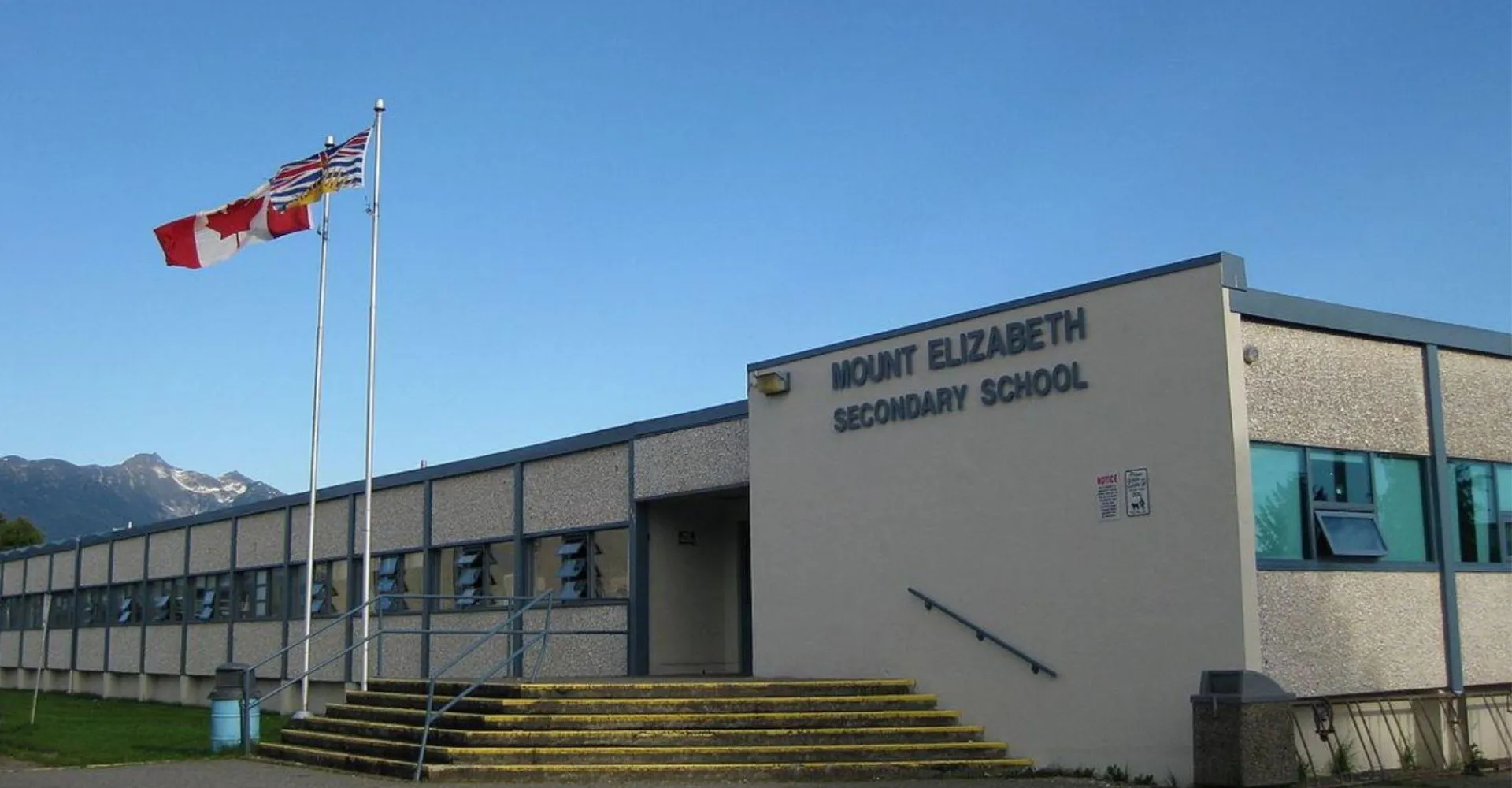 L'edificio scolastico moderno della Mount Elizabeth Middle/Secondary School con la bandiera canadese e americana sotto un cielo limpido.