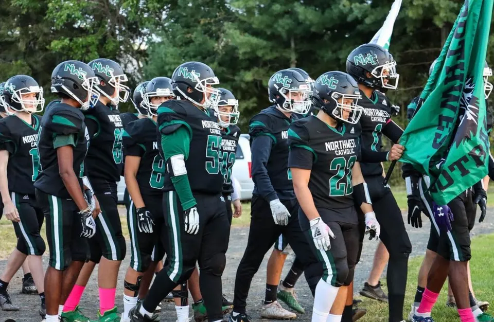 Un gruppo di calciatori della Northwest Catholic High School posa insieme davanti a uno sfondo verde di alberi e piante.