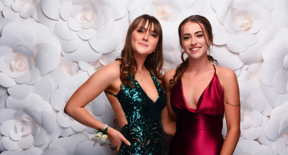 Due studentesse della ACG Strathallan posano in eleganti abiti da sera davanti a uno sfondo di grandi fiori di carta bianchi.