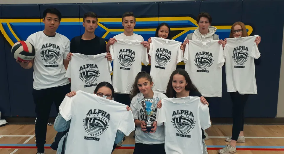 Un gruppo di studenti e studentesse della Alpha School posa in magliette bianche con il logo Alpha in una palestra con pareti blu e gialle.