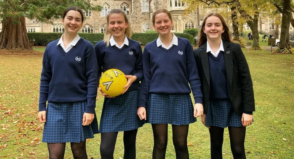Quattro studentesse della Royal High School in divisa scolastica si trovano su un prato e una di loro tiene un pallone giallo.