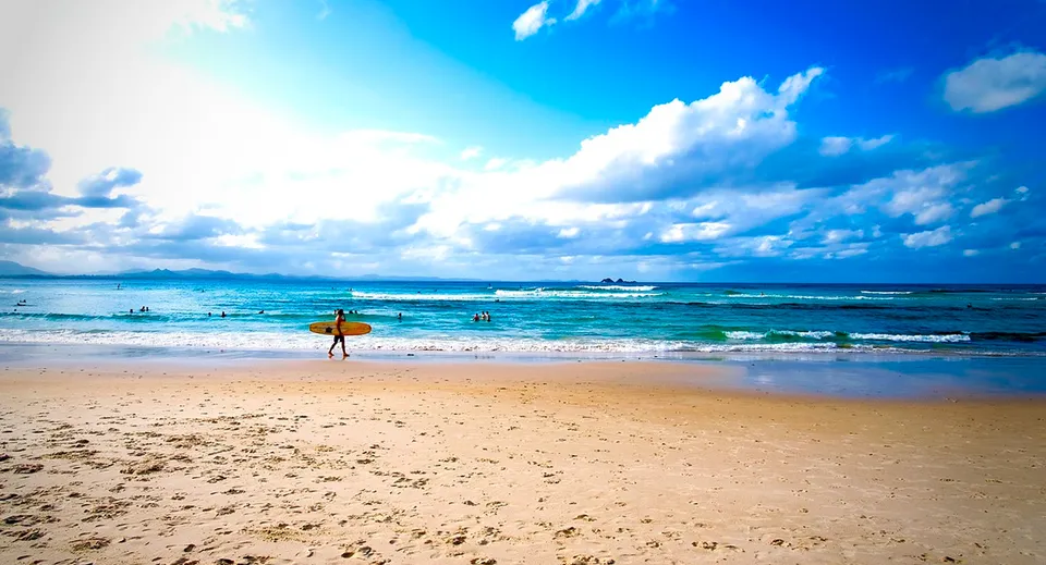 Un surfer cammina lungo la spiaggia soleggiata sotto un cielo blu nell'area della Eastern Fleurieu R-12 School.