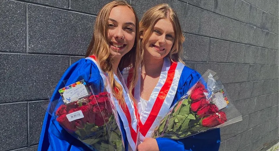Due diplomande in toghe colorate con mazzi di fiori davanti a una parete di cemento grigio alla Bear Creek Secondary School.