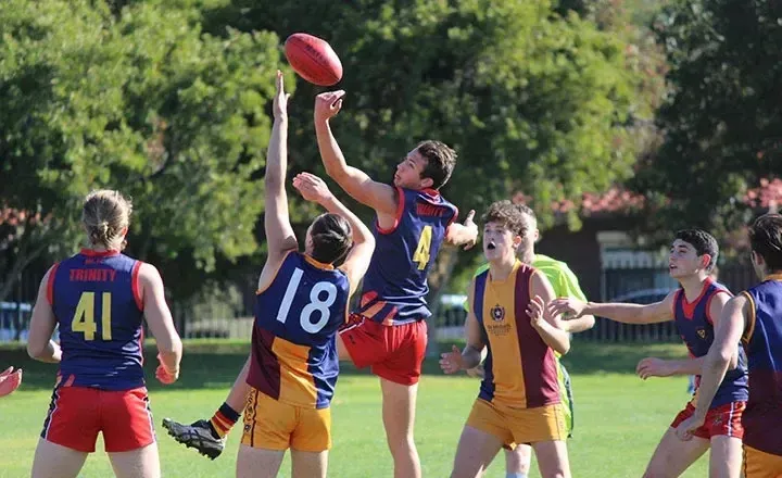 Estudiantes del Trinity College juegan al fútbol australiano en un campo de césped rodeado de vegetación.