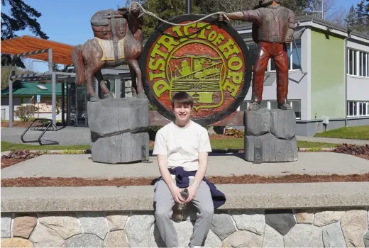 Stefan sitzt vor dem Schild „District of Hope“ mit Holzskulpturen in Kanada