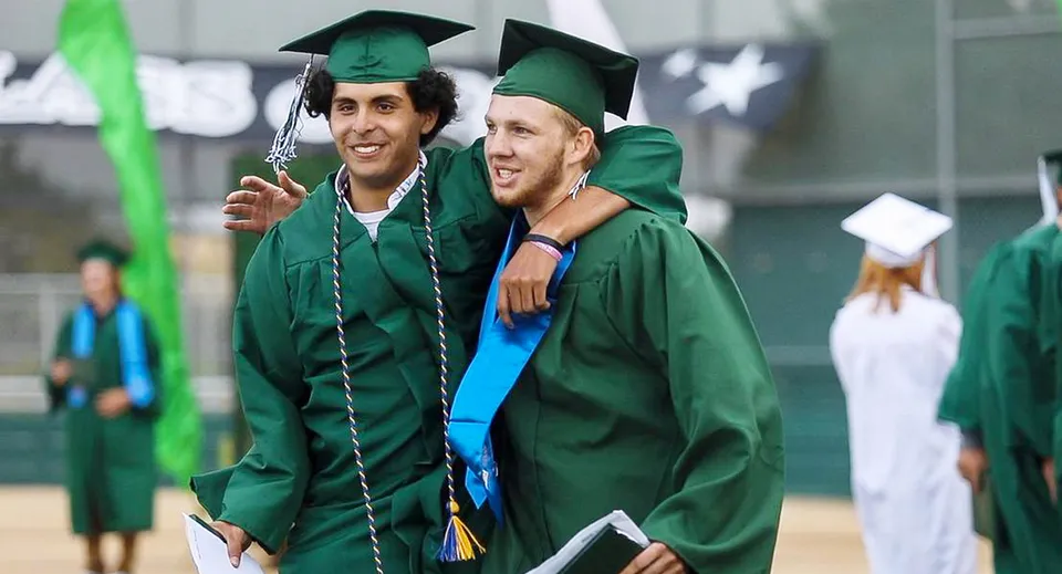Due diplomati della Templeton Secondary School si abbracciano in toga verde davanti ad altri diplomati.