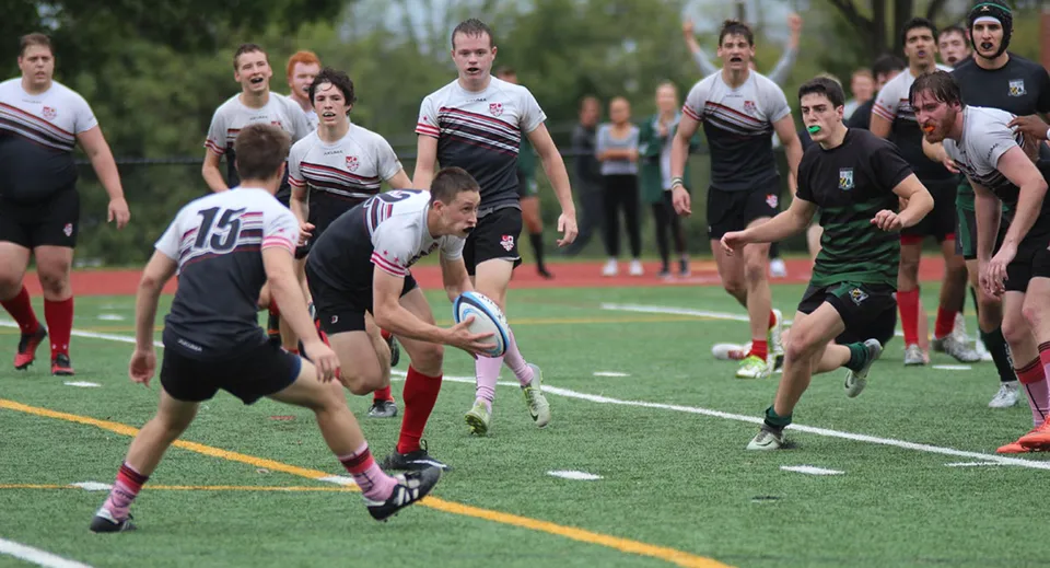 Studenti della Catholic University School giocano a rugby su un campo d'erba con spettatori sullo sfondo.