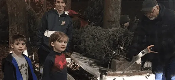 Un hombre corta la base de un pino navideño con serrucho mientras varios niños observan