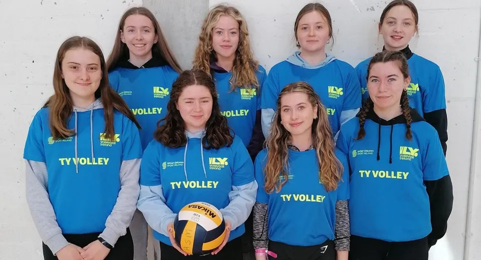 Un gruppo di giovani giocatrici di pallavolo in maglia blu posa davanti a una parete bianca nel campus della Ardscoil Mhuire.