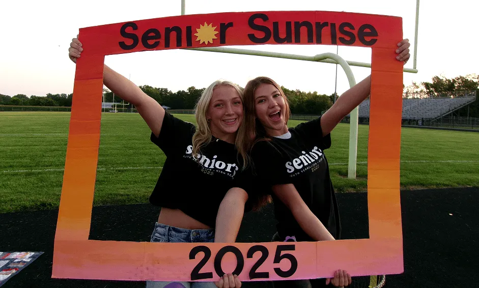 Dos alumnas sujetando un marco de cartón con el lema Senior Sunrise 2025 en el campo del instituto