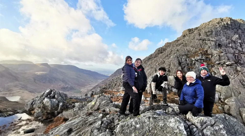 Un gruppo di studenti del Concord College si trova su uno sperone roccioso con montagne e cielo nuvoloso sullo sfondo.