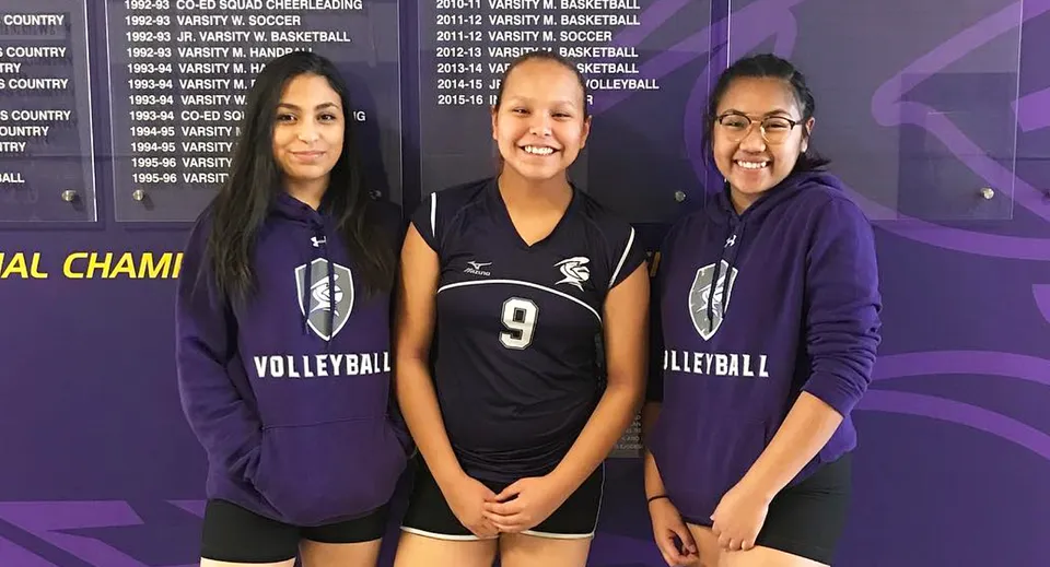 Tre studentesse della Queen Elizabeth High School sono in piedi con le loro divise da pallavolo davanti a una parete con riconoscimenti sportivi.