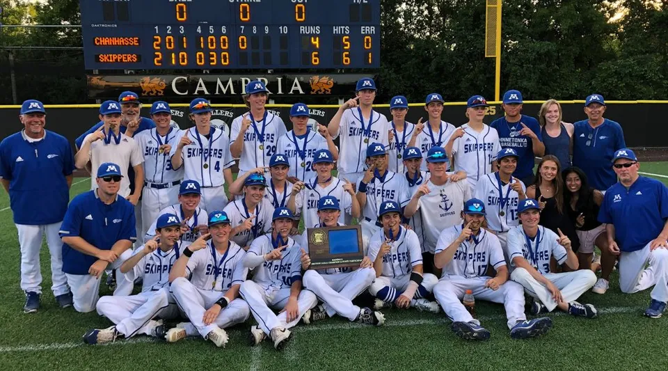 Un gruppo di giocatrici e giocatori di baseball in posa sul campo da baseball delle Minnetonka Public Schools davanti al tabellone segnapunti.
