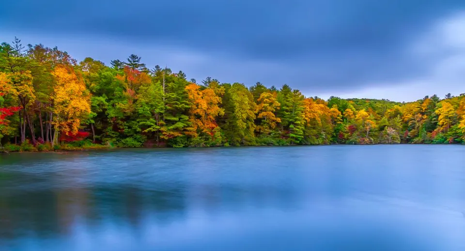Un lago calmo circondato da un bosco autunnale con foglie colorate sullo sfondo del cielo blu.