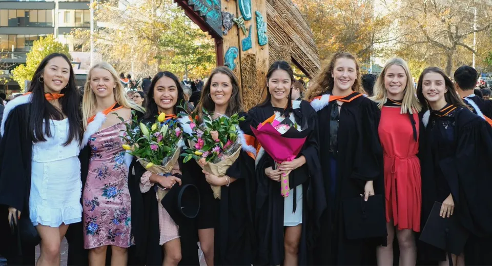 Un gruppo di studentesse diplomate della Onehunga High School si trova davanti a un murale colorato tenendo in mano dei mazzi di fiori.