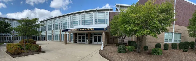 Das moderne Schulgebäude der Chelsea High School mit einer Glasfassade und umgeben von Bäumen und Grünflächen.