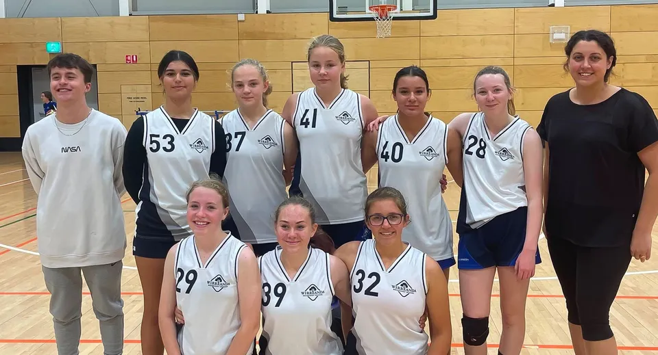 Un gruppo di giovani giocatrici di basket posa insieme nella palestra della Wirreanda Secondary School davanti a un canestro.