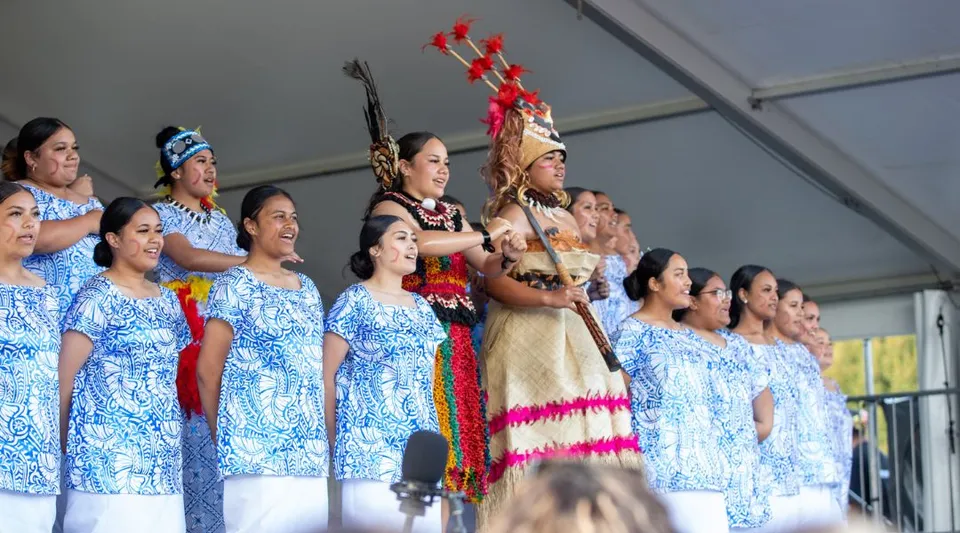 Studentesse della Avonside Girls' High School si esibiscono in abiti tradizionali hawaiani su un palco davanti a una parete grigia.