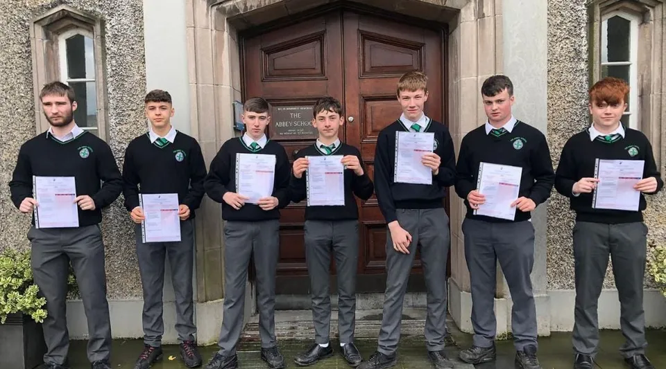 Un gruppo di studenti e studentesse della The Abbey School è in piedi davanti a un grande portone di legno e tiene in alto dei certificati.