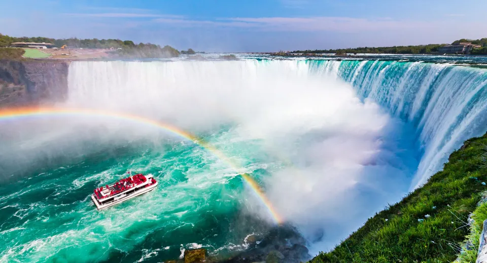 Un'imbarcazione turistica naviga sull'acqua turchese sotto un arcobaleno davanti alle Cascate del Niagara.